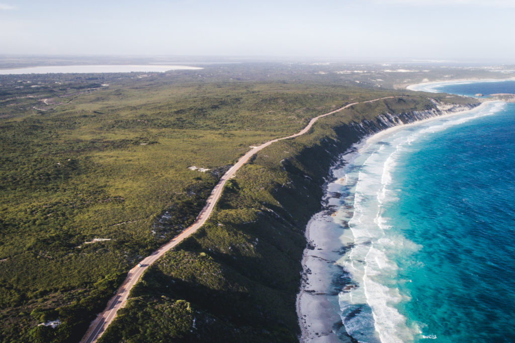 A drone shot of a long stretch of road next to pristine blue coastline