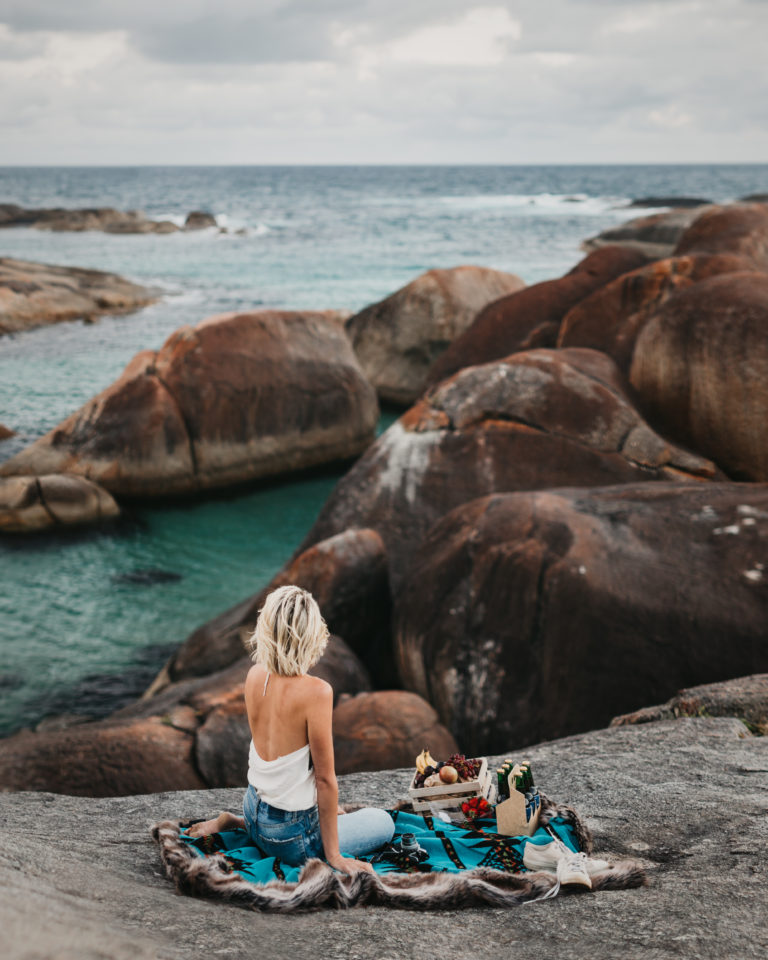 A woman sits on a rug enjoying a picnic on giant boulders facing the ocean