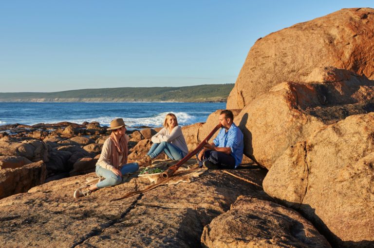 An image of three people sitting on coastal rocks while an Indigenous man plays the digeridoo to show the cultural experiences had along The South West Edge road trip