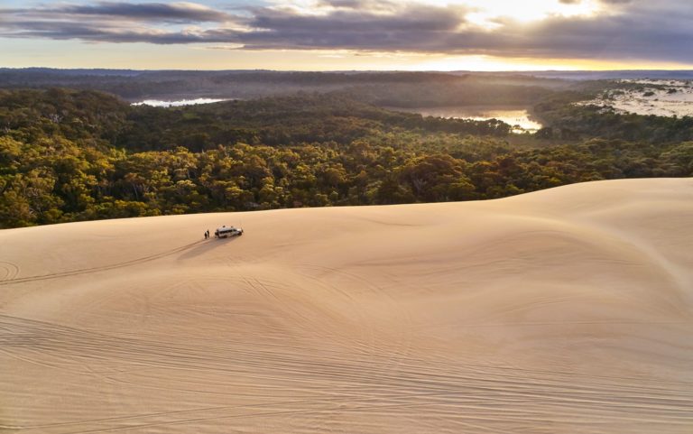 An aerial image of people and a four wheel drive vehicle on huge sand dunes overlooking forest and water shows impressive natural landscapes found on The South West Edge road trip