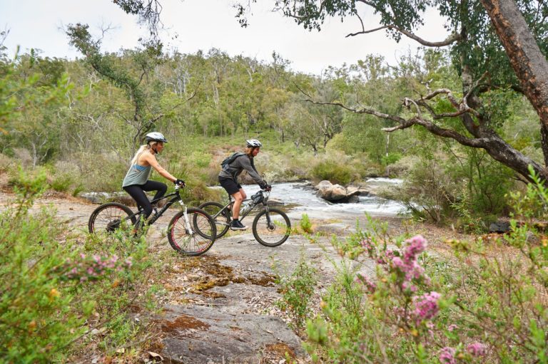 An image of a couple riding mountain bikes in a forest with river beside them shows outdoor adventure experiences immersed in nature while on The South West Edge road trip