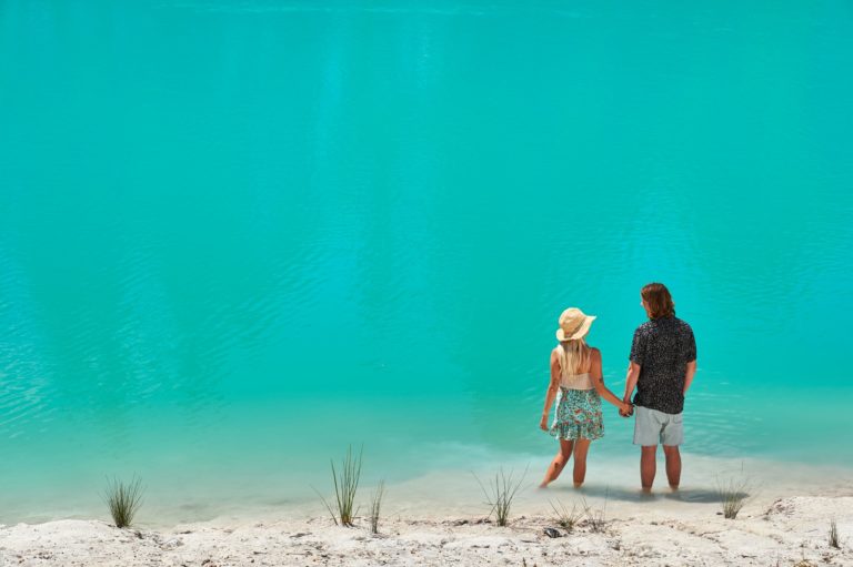 An image of a couple holding hands on the banks of a turquoise blue lake to show amazing natural attractions en route along The Edge south western australia road trip