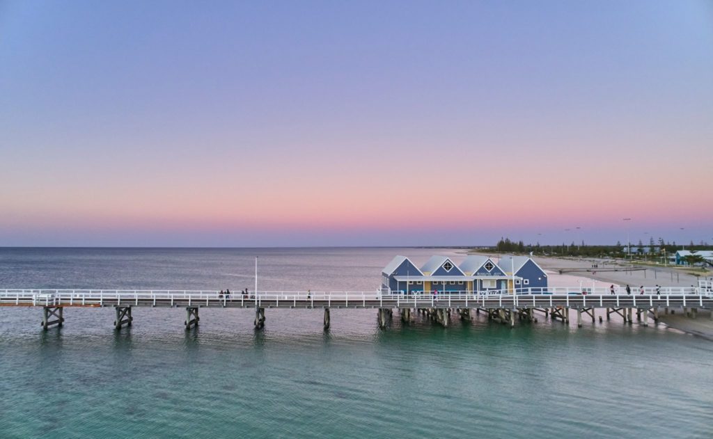 An aerial image shows four blue boat-houses a the beginning of Busselton Jetty with pastel pink and purple sunset sky to show the beautiful tourist attractions on The South West Edge road trip