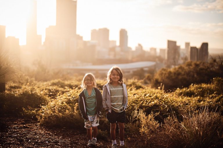 Two kids smile from a garden with Perth City behind them to show the city is a great place for a family holiday