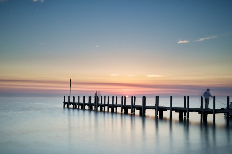A pastel pink blue and yellow sunrise over calm ocean and a boat ramp shows simple beauty found along this road trip down south western australia