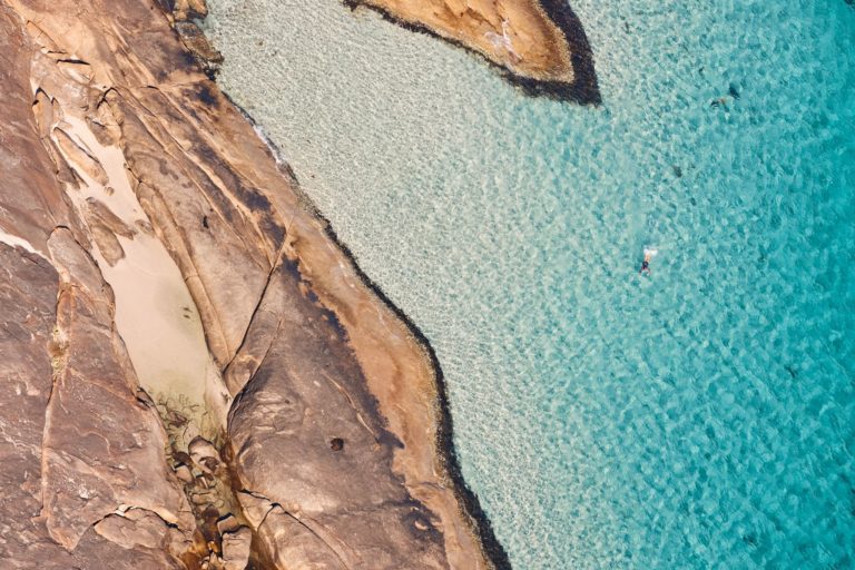 A drone image of a lone swimmer in crystal blue ocean water to show pristine natural attractions on the south western australia road trip The Edge