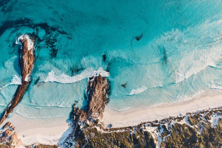 A drone shot of pristine blue ocean with rock formations and white sand to show the incredible natural attractions on The South West Edge road trip