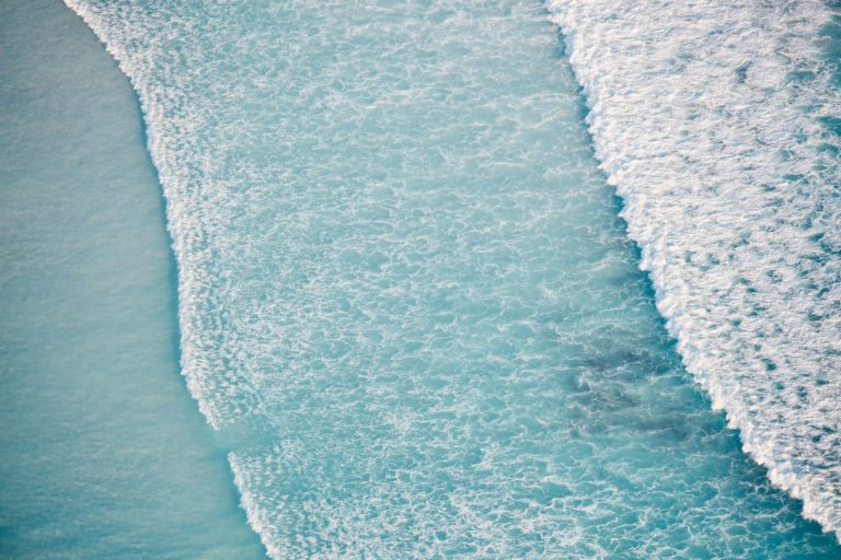 An aerial shot of lapping frothy waves of crystal blue ocean show the untouched natural beauty found on the perth to esperance road trip The Edge