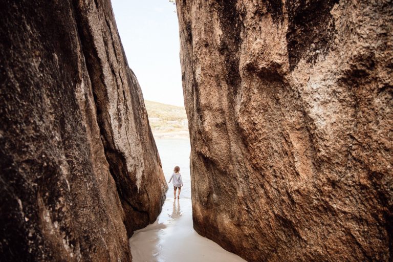 A small child walks through a thin walkway between two granite boulders on a beach to show the rugged natural landscape found on The South West Edge road trip in south west australia