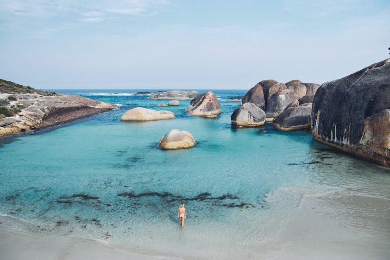 A woman wades in shallow ocean water that is bright peppermint colour with giant boulders to show the incredible landscapes along the esperance to perth road trip of south west australia