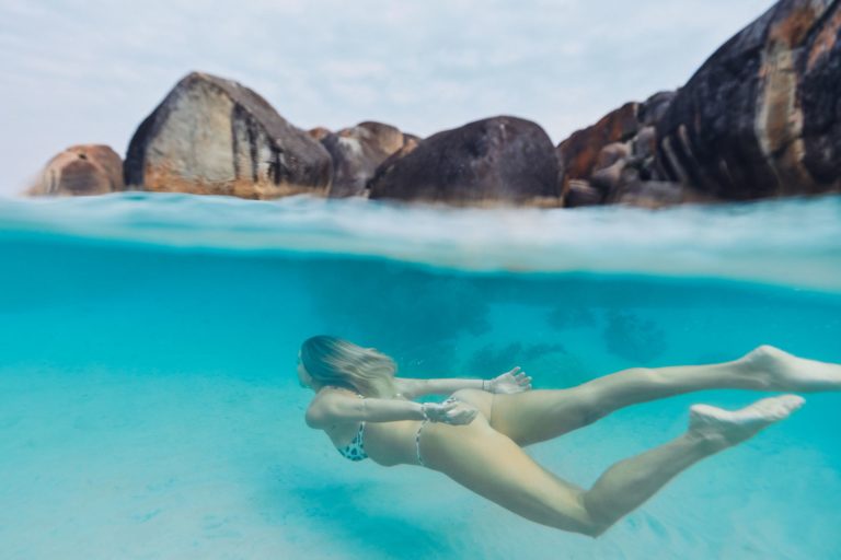 An underwater image of a woman gliding through crystal clear blue waters with boulders in the background to show the incredible natural attractions on the south west australia road trip The Edge