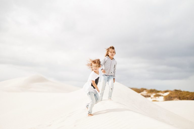 Two children with windswept hair stand on white sand dunes to show incredible natural landscapes to explore along the south western australia road trip The Edge