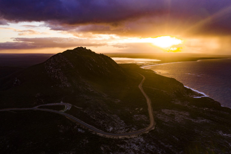 An aerial shot of a mountain surrounded by a winding road with bright yellow and purple sunset shows epic landscapes found on The South West Edge road trip