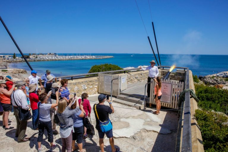 An image shows a tour group taking photos of a canon being fired into blue ocean to show historical experiences and tours while driving en route along The South West Edge road trip