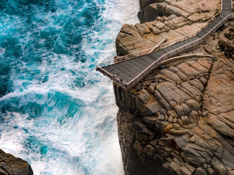 A drone image shows people looking over the edge of the hanging viewing platform at cliffs above the southern ocean to show the rugged landscape and natural monuments on The South West Edge road trip