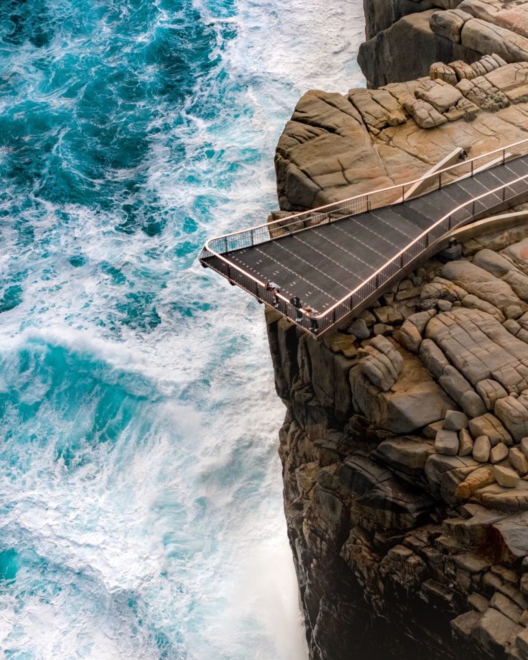 A portrait image of people looking over the edge of a viewing platform that hangs off a cliff over the southern ocean to show the rugged landscape and wild beauty found on the perth to esperance road trip The Edge