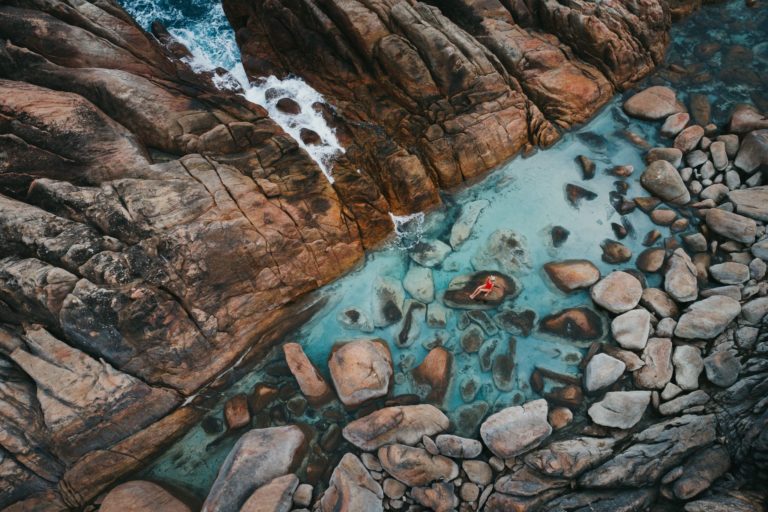 A drone image of a girl in a red swimsuit sitting on a rock with a crystal clear rockpool and mini ocean waterfall shows the unique natural monuments found on The South West Edge road trip
