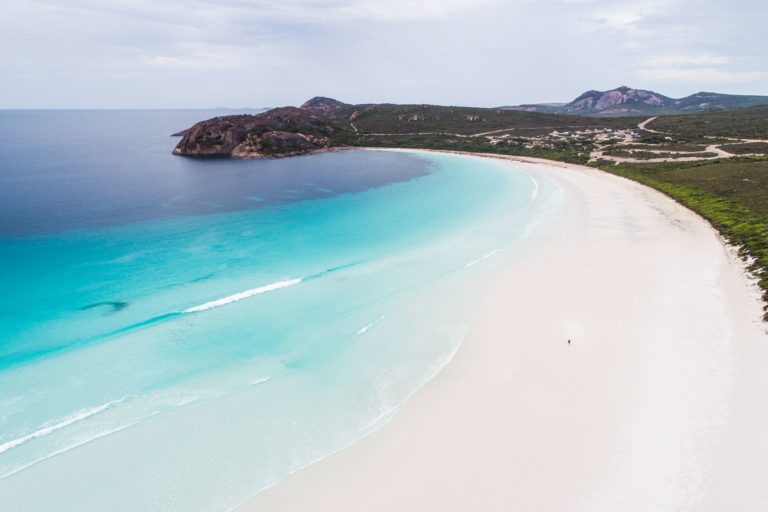A drone photo of a lone person walking on a secluded beach with turquoise water and rugged coastal hills in the distance shows the epic landscapes of The South West Edge road trip