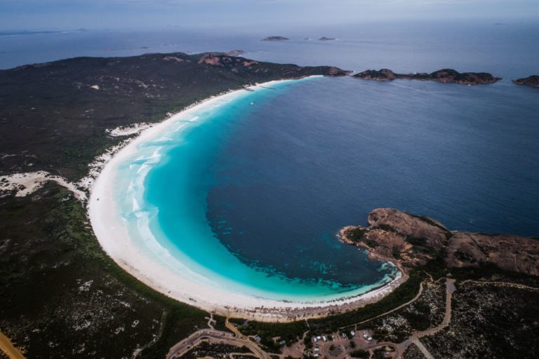 A drone photo of a whole bay with pristine white sand and turquoise blue ocean surrrounded by bushland and roads shows the rugged landscape of the coast along The South West Edge road trip from perth to esperance