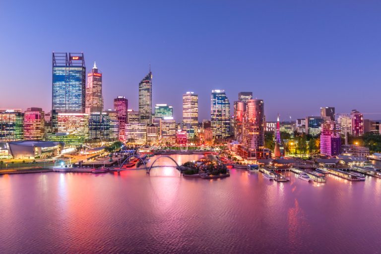 An aerial photo of Perth City lit up at night with pink reflections in the swan river show the bustling city start and end of The South West Edge road trip