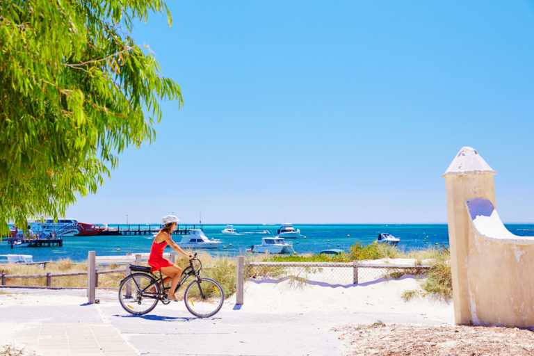 An image of a woman riding a bike with white sand and beach in the background to show outdoor attractions on The South West Edge road trip