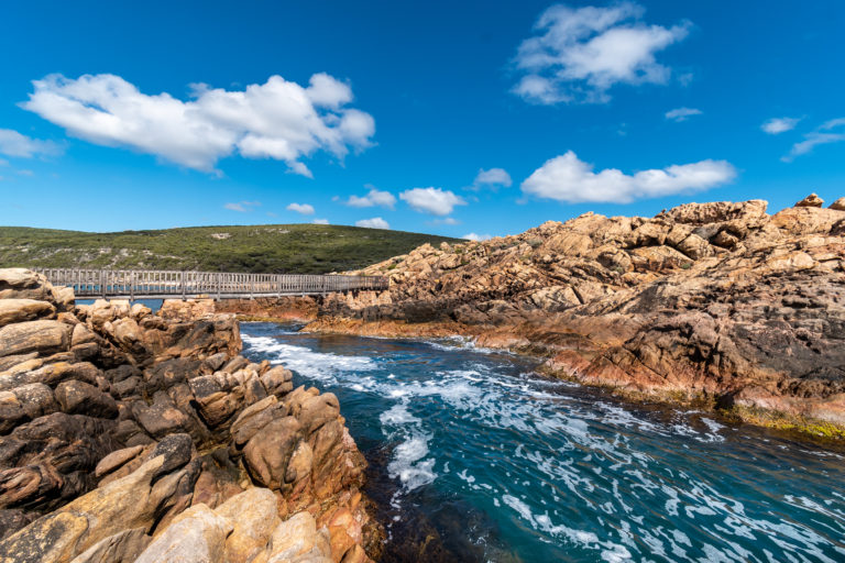 A landscape image of a wooden bridge connecting coastal rock formations with gushing crystal blue water in the middle to show incredible rugged landscapes on The South West Edge road trip
