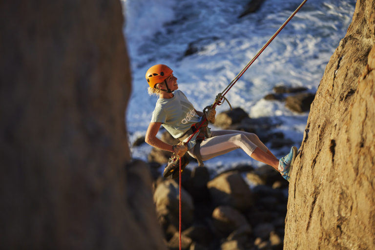 A landscape image of a woman abseiling down a rockface with ocean in the background to show outdoor adventure experiences on The South West Edge road trip