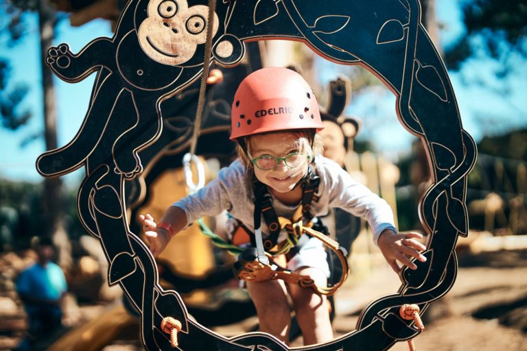 A child wearing an orange helmet peeps through a hole. Shes wearing a harness and glasses