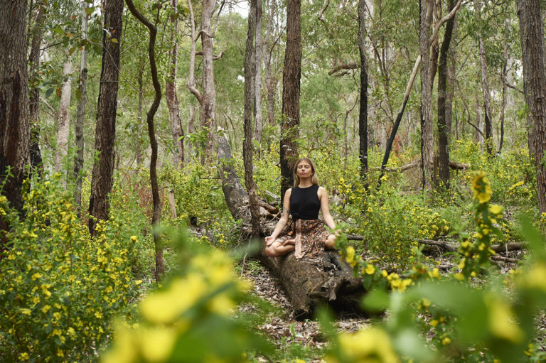 A girl sits cross-legged on a tree trunk surrounded by nature and meditates to show mindfullness along the south west edge road trip