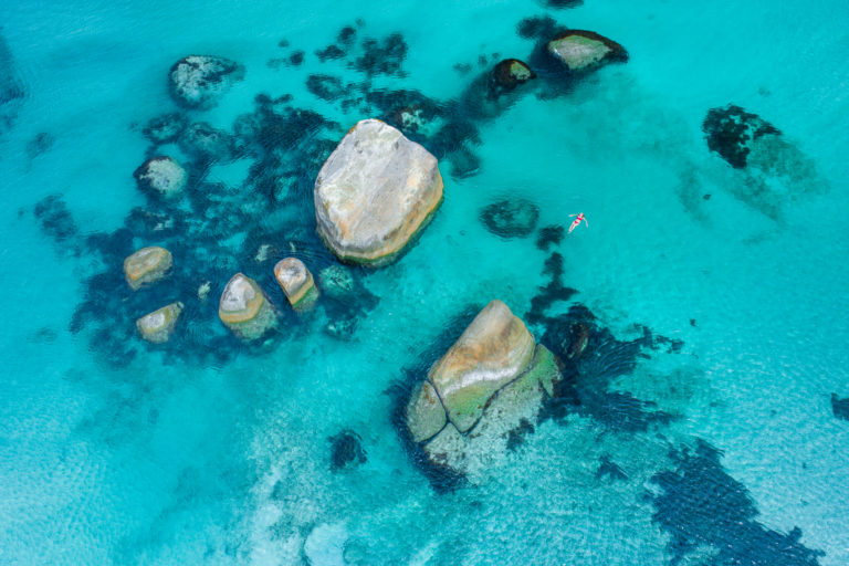 top down drone image of girl in red bikini floating in peppermint coloured waters with giant boulders to show epic landscapes en route along the edge