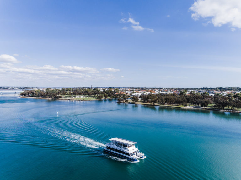 A charter boat cruises on blue water with town in background in Mandurah en route along The Edge