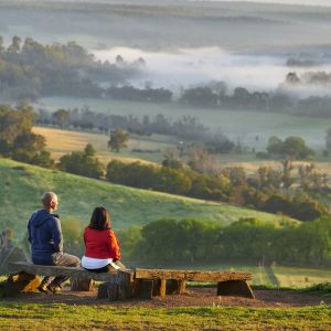 Two people sit on a bench overlooking rolling green hills with mist to show views from Balingup en route along The Edge