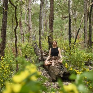 A girl sits cross-legged on a tree trunk surrounded by nature and meditates to show mindfullness along the south west edge road trip