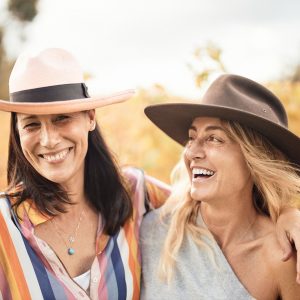 Two women smiling and laughing with a vineyard behind them shows happiness while travelling the south western australia road trip
