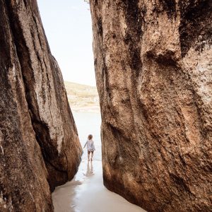 A small child walks through a thin walkway between two granite boulders on a beach to show the rugged natural landscape found on The South West Edge road trip in south west australia
