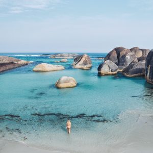 A woman wades in shallow ocean water that is bright peppermint colour with giant boulders to show the incredible landscapes along the esperance to perth road trip of south west australia
