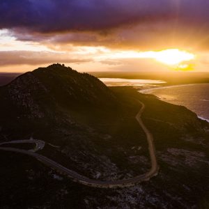An aerial shot of a mountain surrounded by a winding road with bright yellow and purple sunset shows epic landscapes found on The South West Edge road trip