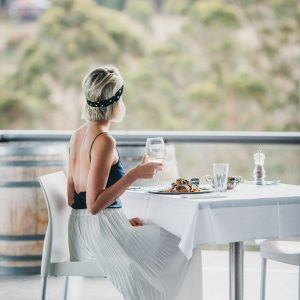 A woman sits at a table overlooking nature and sips on a glass of wine with a gourmet meal to show the eat and drink experiences to be had along The South West Edge road trip