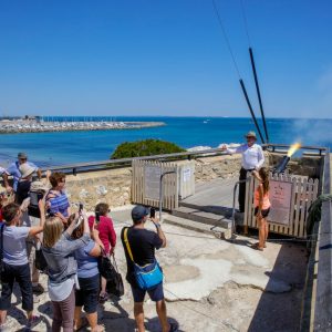 An image shows a tour group taking photos of a canon being fired into blue ocean to show historical experiences and tours while driving en route along The South West Edge road trip