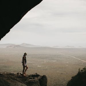 A woman stands in a cliff cave looking at the vast sweeping landscape and a long road to show the natural monuments and wild beauty along The South West Edge road trip