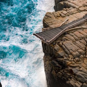 A drone image shows people looking over the edge of the hanging viewing platform at cliffs above the southern ocean to show the rugged landscape and natural monuments on The South West Edge road trip