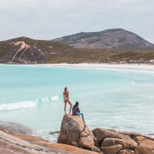 A couple stands on a rock overlooking a pristine beach with rocky hills in the background to show the vivid contrasts of The South West Edge road trip and landscape
