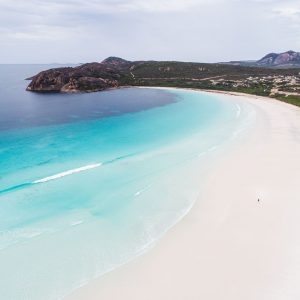 A drone photo of a lone person walking on a secluded beach with turquoise water and rugged coastal hills in the distance shows the epic landscapes of The South West Edge road trip