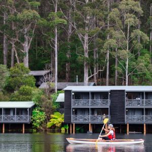 girl kayaks on green lake with chalets and tall karri trees in background shows tranquil accommodation options and activities en route along the south west edge